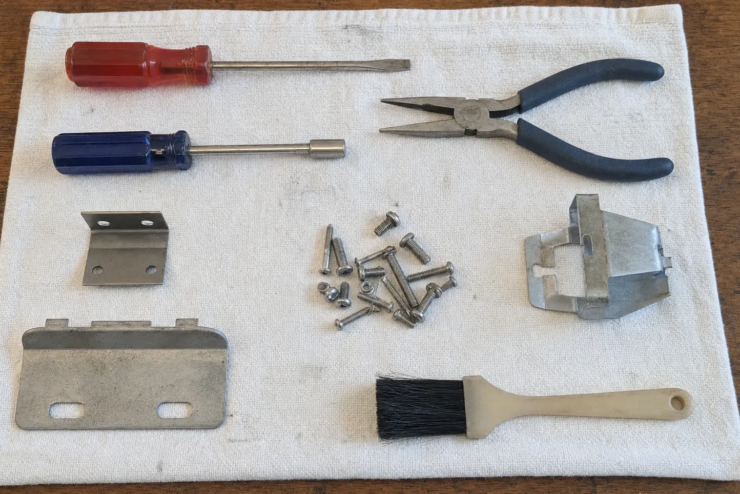 Screwdrivers, needle-nose pliers, a brush, machine screws and two sheet-metal brackets laid out on a cloth